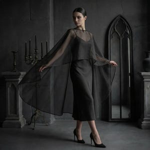 May include: A black dress with a sheer cape. The dress is a midi length with a simple neckline. The cape is long and flowing, with a delicate, transparent fabric. The model is wearing black heels. The background is a dark gray room with gothic-style decor.