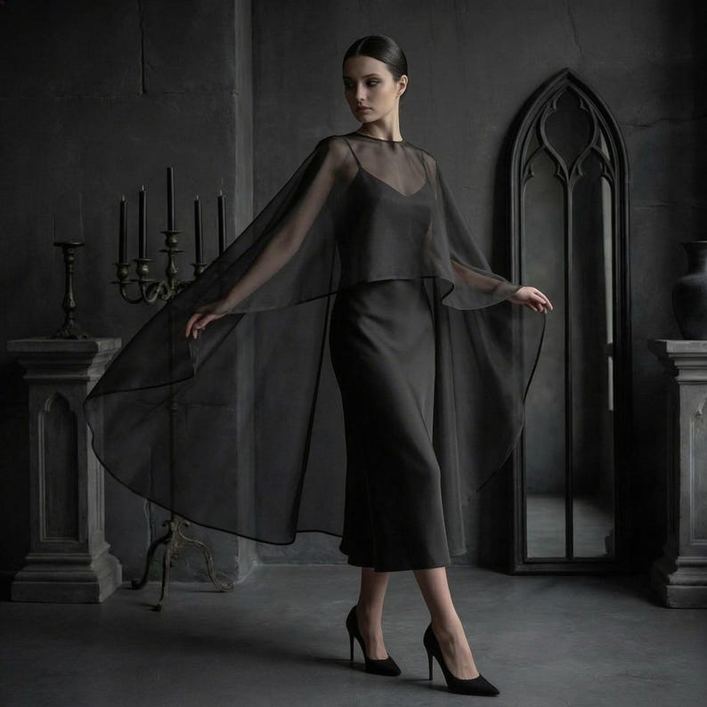 May include: A black dress with a sheer cape. The dress is a midi length with a simple neckline. The cape is long and flowing, with a delicate, transparent fabric. The model is wearing black heels. The background is a dark gray room with gothic-style decor.
