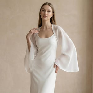 May include: A white, floor-length dress with a scoop neckline and a sheer, flowing bolero. The bolero has wide, bell sleeves. The dress and bolero are made of a lightweight fabric, creating an elegant and airy look.