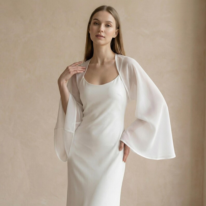 May include: A white, floor-length dress with a scoop neckline and a sheer, flowing bolero. The bolero has wide, bell sleeves. The dress and bolero are made of a lightweight fabric, creating an elegant and airy look.