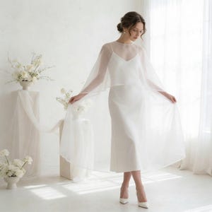 May include: A white wedding dress with a sheer overlay, featuring a V-neckline and spaghetti straps. The dress is paired with white heels. The background includes white floral arrangements and a sheer curtain.