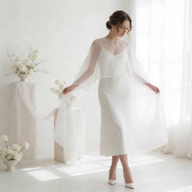 May include: A white wedding dress with a sheer overlay, featuring a V-neckline and spaghetti straps. The dress is paired with white heels. The background includes white floral arrangements and a sheer curtain.