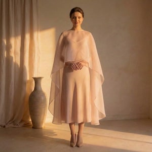 May include: A woman wearing a light peach-colored outfit. The outfit includes a sleeveless top, wide-leg pants, a sheer cape, and matching heels. A large, neutral-toned vase is visible in the background, along with a curtain.