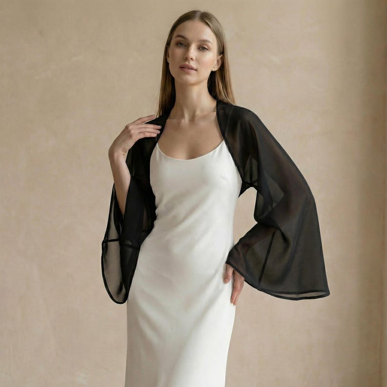 May include: A black sheer bolero with wide sleeves is worn over a white slip dress. The bolero has a delicate, flowing appearance. The woman is standing in front of a neutral-toned background.