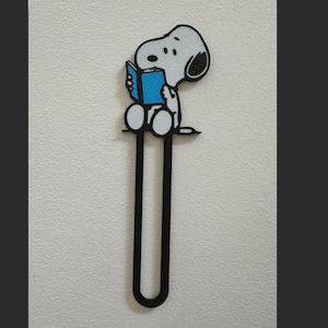Può includere: Un segnalibro nero con Snoopy dei Peanuts, che legge un libro blu. Snoopy è bianco con orecchie e macchie nere. Il segnalibro ha un lungo design a forma di U.