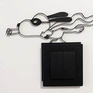 May include: A black metal wall art piece depicting a cartoon dog in a superhero pose, complete with a cape and goggles. The art is mounted above a black light switch with two buttons, creating a playful contrast against the white wall.