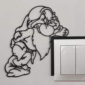 Può includere: Arte da parete in metallo nero di Brontolo di Biancaneve, appoggiato a un interruttore della luce bianco. L'opera d'arte è una silhouette, che mostra i tratti distintivi di Brontolo, tra cui la sua barba e il suo cappello. L'interruttore ha due pulsanti rettangolari.