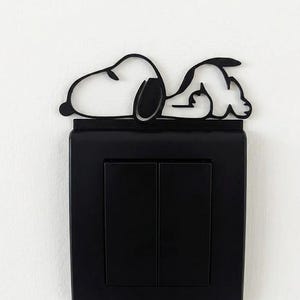 Puede incluir: Una cubierta de interruptor de luz negra con un diseño de Snoopy. La silueta de Snoopy está acostada encima de la placa del interruptor. La placa del interruptor tiene dos interruptores rectangulares. El fondo es una pared blanca lisa.