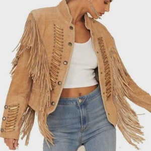 May include: A tan suede jacket with long fringe detailing on the sleeves and chest. The jacket features decorative stitching and button closures. The jacket is worn with a white top and blue jeans.