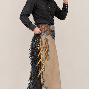 May include: Tan leather chaps featuring intricate blue and metallic gold floral tooling along the edges, accented with long black and blue fringe. The chaps include three vertical orange leather straps and are worn over blue denim jeans.