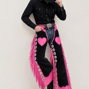 May include: Black suede chaps featuring bright pink heart-shaped patches on the thighs, long pink fringe along the outer legs, and silver-toned metal concho accents. The chaps are worn over blue denim jeans with a black shirt, hat, and boots.