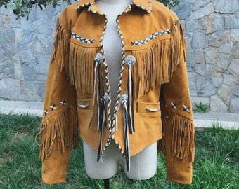 May include: Mustard-colored suede jacket with fringe detailing. The jacket features decorative stitching, silver-toned conchos, and long fringe along the sleeves and chest. The jacket is displayed on a mannequin in a grassy outdoor setting.