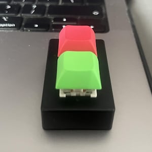 Może przedstawiać: Czarny, prostokątny tester klawiszy klawiatury z dwoma nakładkami na klawisze. Górna nakładka na klawisz jest czerwona, a dolna zielona. Nakładki na klawisze są kwadratowe i osadzone na białej podstawie.