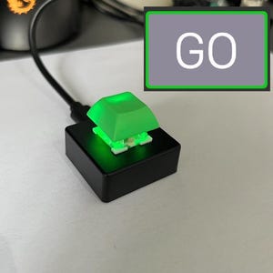 Puede incluir: Un interruptor de teclado mecánico montado sobre una base cuadrada negra con una tecla retroiluminada en verde brillante. Un cable negro sale de la base. Un gráfico rectangular con borde verde contiene el texto blanco GO sobre un fondo gris.