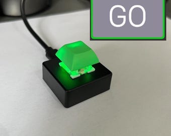 Puede incluir: Un interruptor de teclado mecánico montado sobre una base cuadrada negra con una tecla retroiluminada en verde brillante. Un cable negro sale de la base. Un gráfico rectangular con borde verde contiene el texto blanco GO sobre un fondo gris.