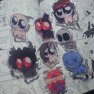 May include: A selection of cartoon character acrylic charms. Each charm has a unique design with large eyes and various colours, including red, blue, and pink. The charms are displayed on a comic book page.