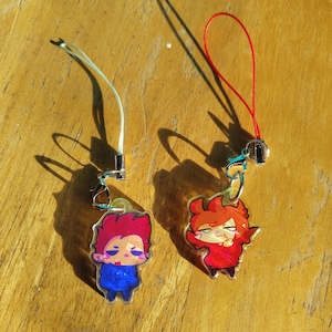 Könnte beinhalten: Zwei Cartoon-Charms mit Lanyards. Ein Charm zeigt einen Charakter mit roten Haaren und einem blauen Outfit. Der andere Charm hat einen Charakter mit roten Haaren und einem roten Outfit. Beide Charms haben einen silberfarbenen Verschluss.