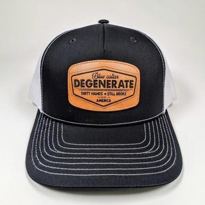 May include: Black and white trucker cap with a brown leather patch. The patch reads "DEGENERATE" above "Blue collar" and "Dirty hands still broke America" in white text. The cap has a curved peak with white stitching.