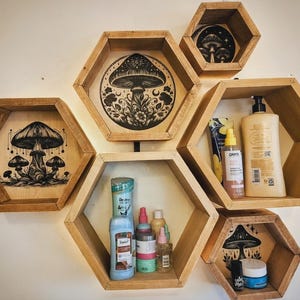 Könnte beinhalten: Sechs hölzerne, sechseckige Regale an einer weißen Wand. Jedes Regal zeigt eine schwarz-weiße Pilzillustration. Die Regale beherbergen verschiedene Toilettenartikel und Kosmetikprodukte.