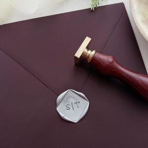 Può includere: Una busta bordeaux sigillata con un sigillo di cera argentato. Il timbro presenta le iniziali S|T all'interno di un rombo. Un timbro di cera con manico in legno è posizionato vicino. La scena è ambientata su una superficie bianca con vegetazione.