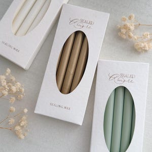 Peut inclure: Trois boîtes de bâtons de cire à cacheter ivoire, dorés et vert menthe. Chaque boîte a une fenêtre ovale affichant les bâtons de cire. Les boîtes sont blanches avec les mots "SEALED Amore" et "SEALING WAX" imprimés.