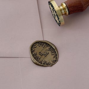 May include: A brass wax seal stamp with a wooden handle rests on a light pink envelope. The stamp has a floral border and the initials "SM" in the center. A matching wax seal is also present.