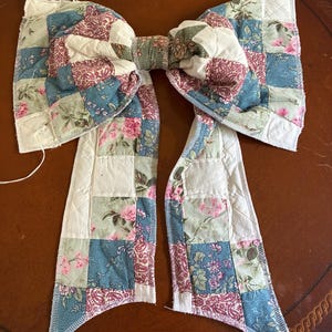 May include: A large decorative bow constructed from quilted fabric patches. The bow features a patchwork design with alternating squares of floral patterns, paisley prints, and solid white fabric in shades of blue, pink, green, and cream.