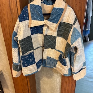 May include: A quilted jacket featuring a patchwork design of various blue and cream fabric squares. The garment has a wide collar, long sleeves with folded cuffs, and three dark buttons down the front. Two patch pockets are visible on the front.