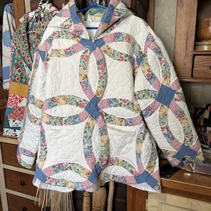 May include: A quilted jacket featuring a white background with a multi-colored floral double wedding ring pattern in shades of pink, blue, green, and yellow. The jacket includes a hood, long sleeves, and two front pockets with a blue fabric trim.