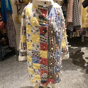 May include: A long-sleeved coat constructed from a patchwork of various fabrics, including yellow, floral, and diamond-patterned squares. The garment features a white collar, a button-front closure, and small white tufted accents throughout.