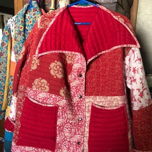 May include: A quilted jacket featuring a patchwork design in various shades of red and cream. The garment has a wide collar, long sleeves, two large front pockets, and a front button closure with four visible dark metal snap buttons.