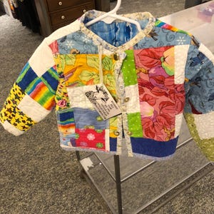 May include: A quilted jacket made from various colorful fabric patches including floral, star, and rainbow patterns. The jacket features a round neckline, long sleeves, and gold-tone metal snap buttons. A white tag with a sheep illustration and text is attached.