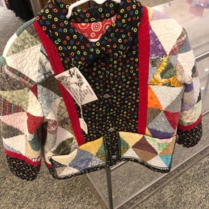 May include: A quilted jacket featuring a patchwork design of various fabric patterns in shades of red, blue, green, and yellow. The jacket has a black collar and front placket with a colorful ring pattern and a tag reading Mulberry Hill Farm.