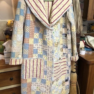 May include: A long-sleeved quilted coat featuring a patchwork design of various light blue, yellow, and white patterned fabric squares. The coat includes a wide shawl collar and two front pockets, both accented with red and white vertical stripes.