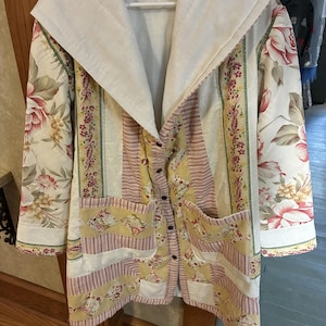 May include: A long-sleeved jacket featuring a patchwork design with floral patterns in pink, yellow, and green, combined with striped panels. The garment includes a large white hood, a button-down front closure, and two front patch pockets.