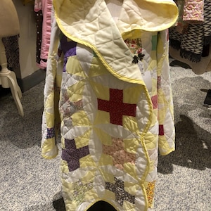 May include: A hooded coat constructed from a patchwork quilt featuring a yellow, white, and multi-colored fabric pattern. The garment has long sleeves, a wide hood, and a quilted texture with various geometric shapes and floral prints.
