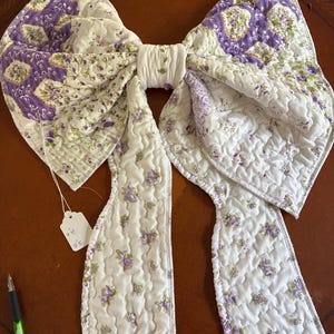 May include: A large quilted fabric bow featuring a white base with purple floral patterns and green vine accents. The bow has two long hanging tails and a central knot. A small white paper tag is attached, showing the text 3" x 15".