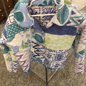 May include: A quilted jacket featuring a patchwork design with various floral, leaf, and geometric patterns in shades of blue, green, white, and purple. The jacket has a wide collar, long sleeves, and a front button closure with a white tag attached.