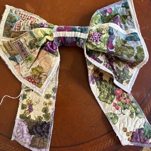May include: A fabric bow featuring a quilted pattern of grapes, vines, and wine labels with text including "Jordana" and "EAU DE L'EAU". The bow has raw, serged edges and a central fabric band, showcasing a multi-colored print of purple and green grapes.