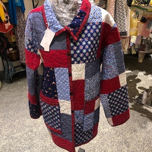 May include: A long-sleeved quilted jacket featuring a patchwork design of various fabric squares in shades of red, white, and blue. The jacket has a collared neckline, a button-down front closure, and a white rectangular price tag attached.
