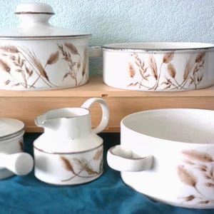 May include: A set of white ceramic kitchenware featuring a brown wheat stalk design. The collection includes a lidded pot, a serving dish, a creamer jug, and a bowl. The items are displayed on a wooden box and a blue cloth.