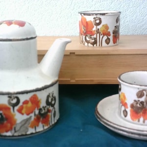 Op de afbeelding: Een vintage keramische theeset met een theepot, twee kopjes en een schotel. De set heeft een crèmekleurige basis met een bloemmotief in oranje, bruin en zwart. De theepot is ongeveer 20 cm hoog.