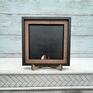 May include: A square wooden picture frame with a black interior and a brown border. The frame is supported by a small wooden easel. The frame is designed to hold a small picture or artwork. The frame is made of wood and has a simple, modern design.