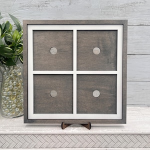 May include: A square, wooden decorative frame with a white grid design, featuring four sections, each with a small, circular accent. The frame is supported by a small, brown wooden stand. The wood has a gray stain.