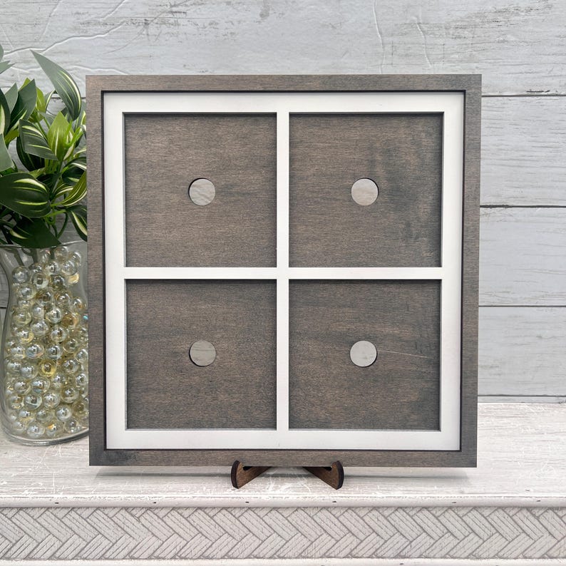 May include: A square, wooden decorative frame with a white grid design, featuring four sections, each with a small, circular accent. The frame is supported by a small, brown wooden stand. The wood has a gray stain.