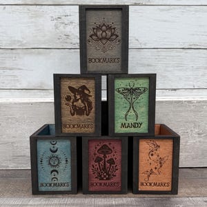 May include: A pyramid stack of six rectangular wooden boxes with dark frames. Each box features a unique laser-engraved design on its front panel, including a lotus, witch, moth, sun and moon, mushrooms, and dragons, with text reading BOOKMARKS or MANDY.