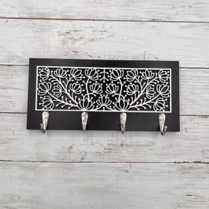 May include: A black wooden key holder with a white floral design and four silver hooks. The decorative panel features a cut-out pattern of stylized flowers and vines. The key holder is mounted on a light-colored wooden background.