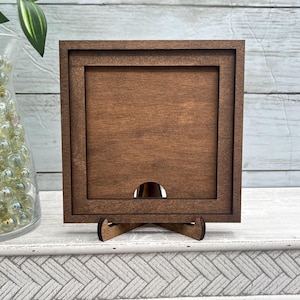 May include: A square, wooden picture frame with a dark brown finish. The frame has a recessed center and a small, arched opening at the bottom. It stands on a matching wooden easel, perfect for displaying photos or artwork.