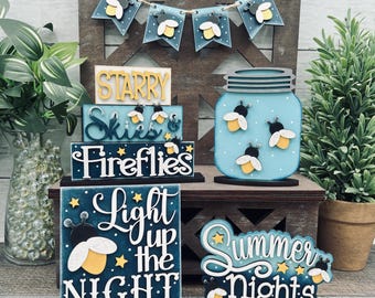 May include: A collection of wooden decorative items with a firefly theme. Includes a banner, blocks with text like "Starry Skies" and "Fireflies", a mason jar, and a sign that says "Light up the Night". The color scheme is blue, white, yellow, and black.