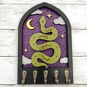 May include: A black wooden key holder with a gothic arch design. It features a green snake with leaf accents against a purple starry background. Includes a crescent moon, clouds, stars, and five brass hooks.